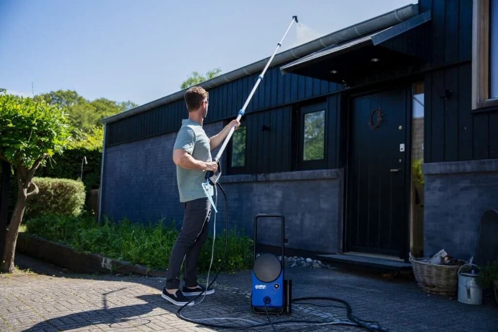 En mann bruker en Nilfisk-teleskopstang med høytrykksdyse for å rengjøre et tak med svarte fliser. Han står foran et hus med lyse vegger og store vinduer.